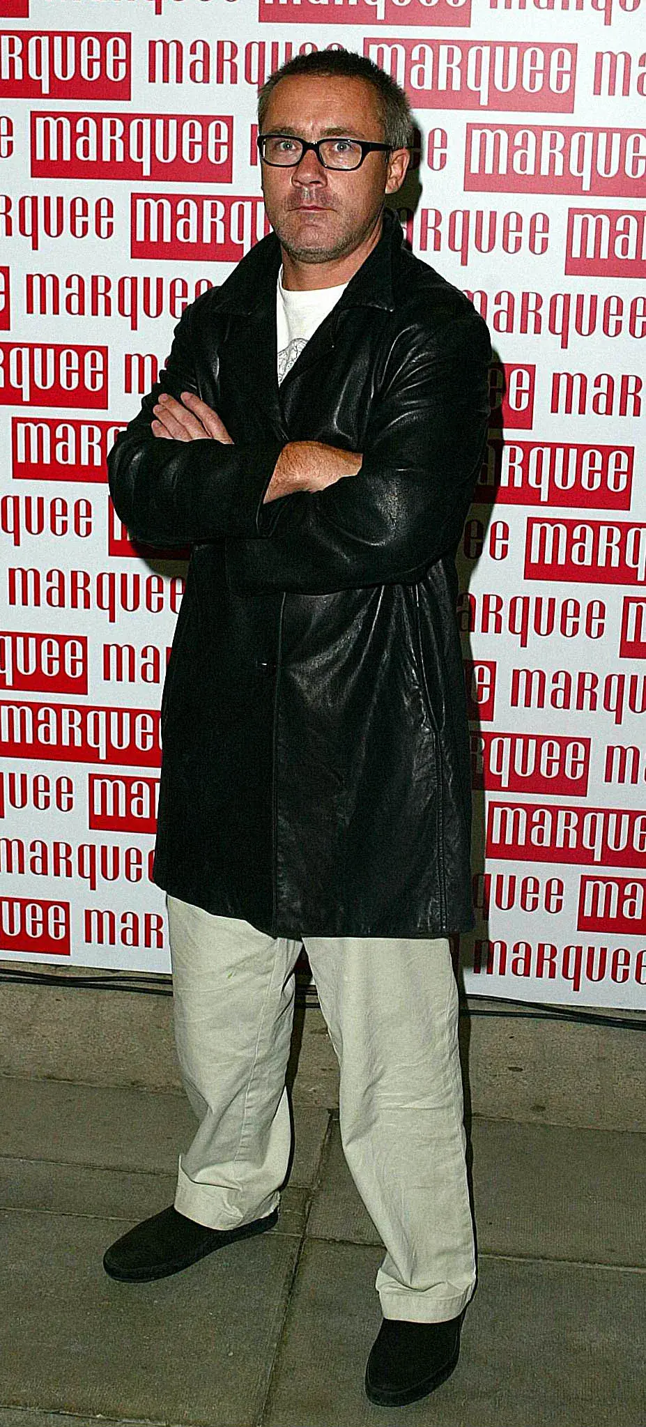 Damien Hirst standing with arms folded in front of Marquee Club branding in 2002