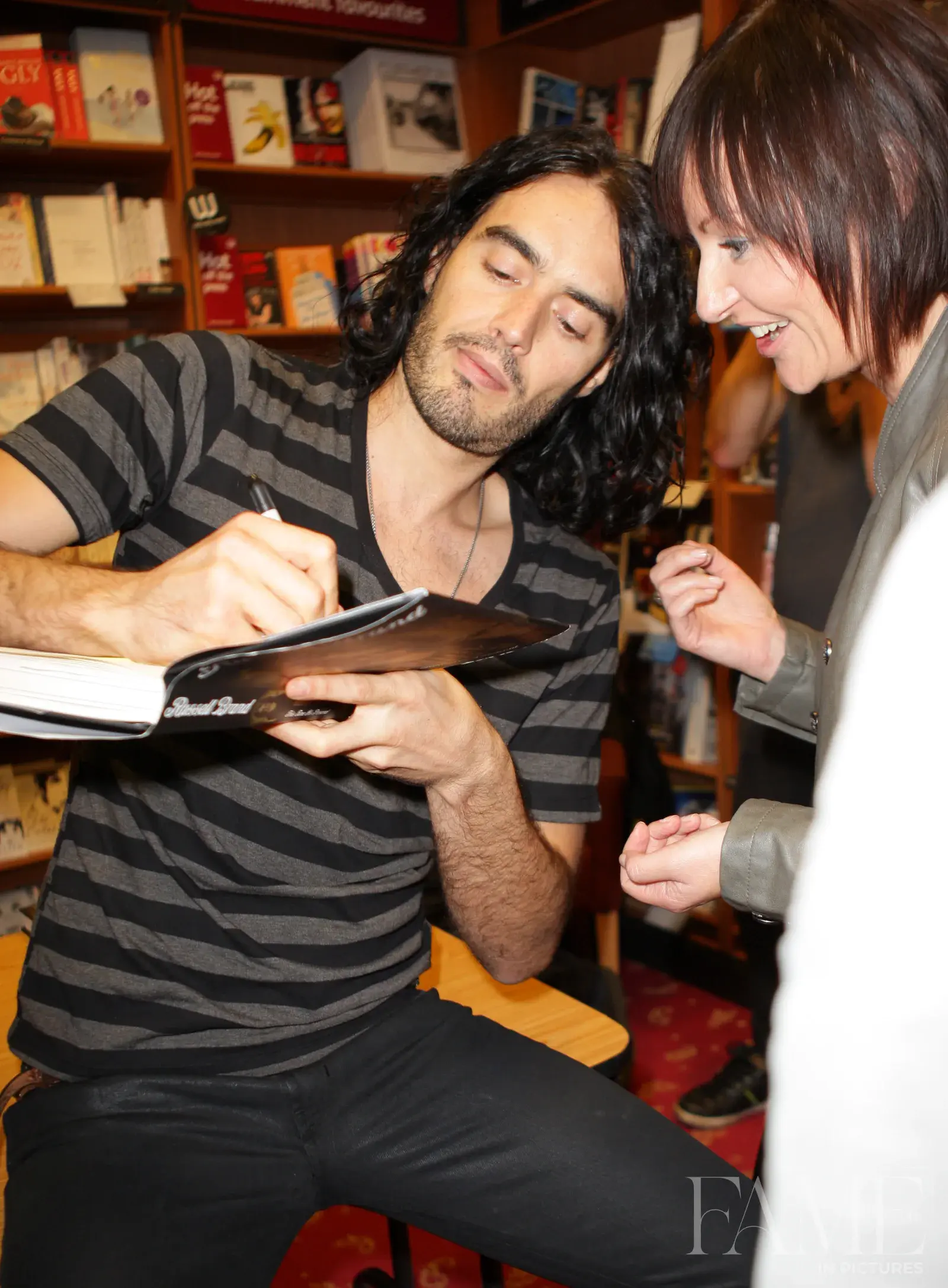 Russell Brand seated at a book signing, writing in a copy while an attendee stands beside him.