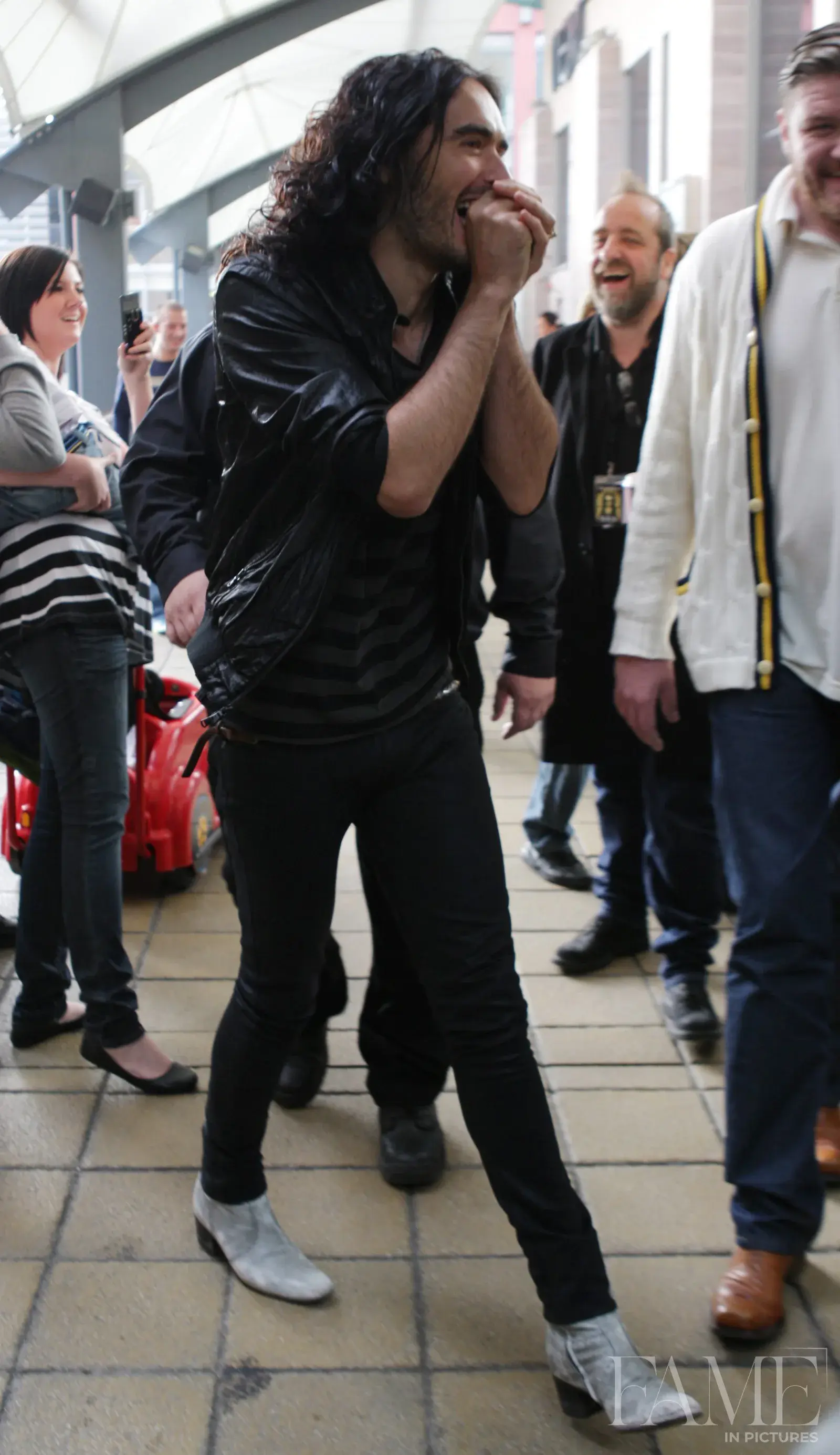 Russell Brand standing in the Merry Hill shopping centre in a dark jacket, striped top and light-coloured boots.