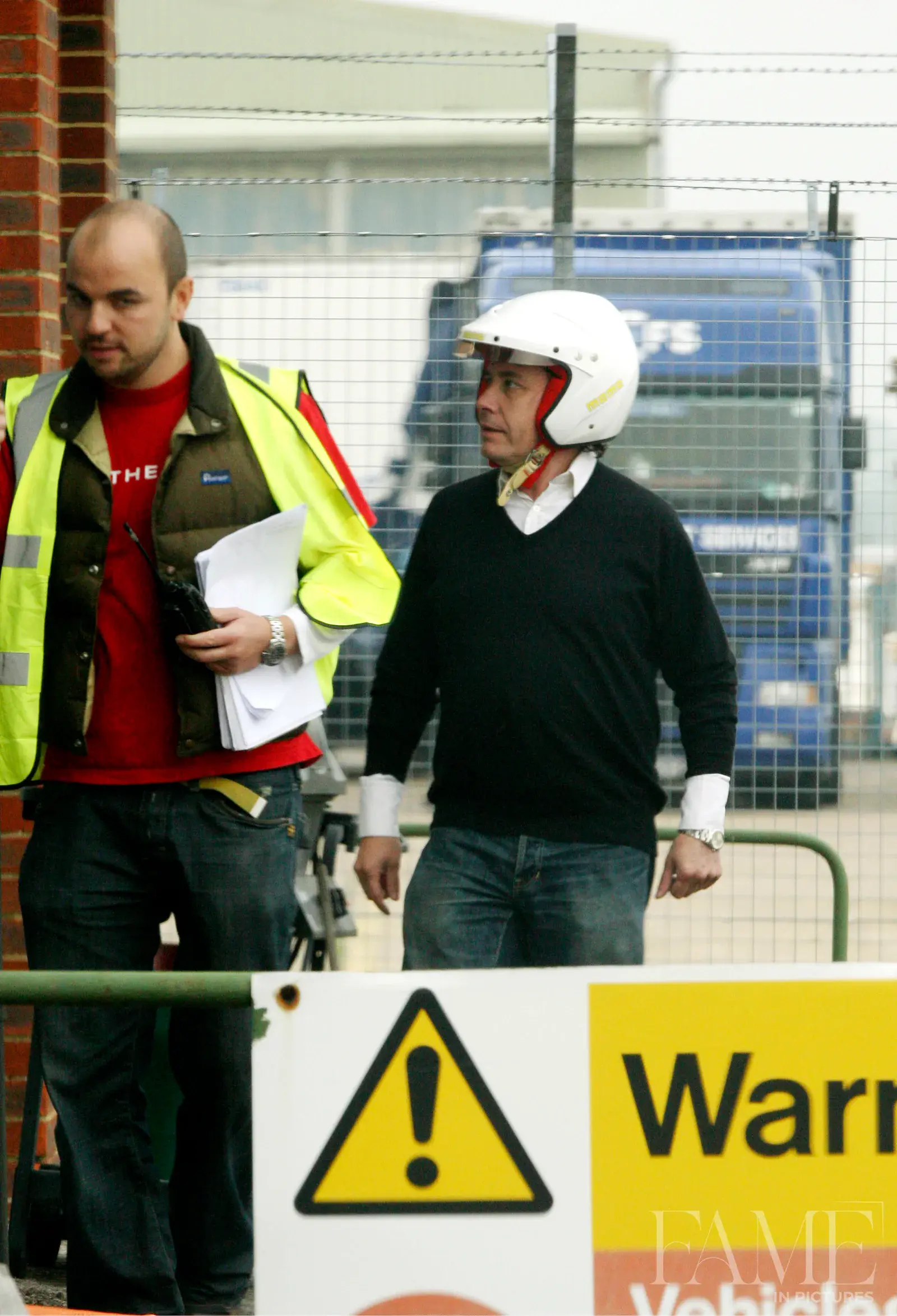 Jools Holland wearing an open-face white helmet and dark jumper beside a man in a high-visibility jacket