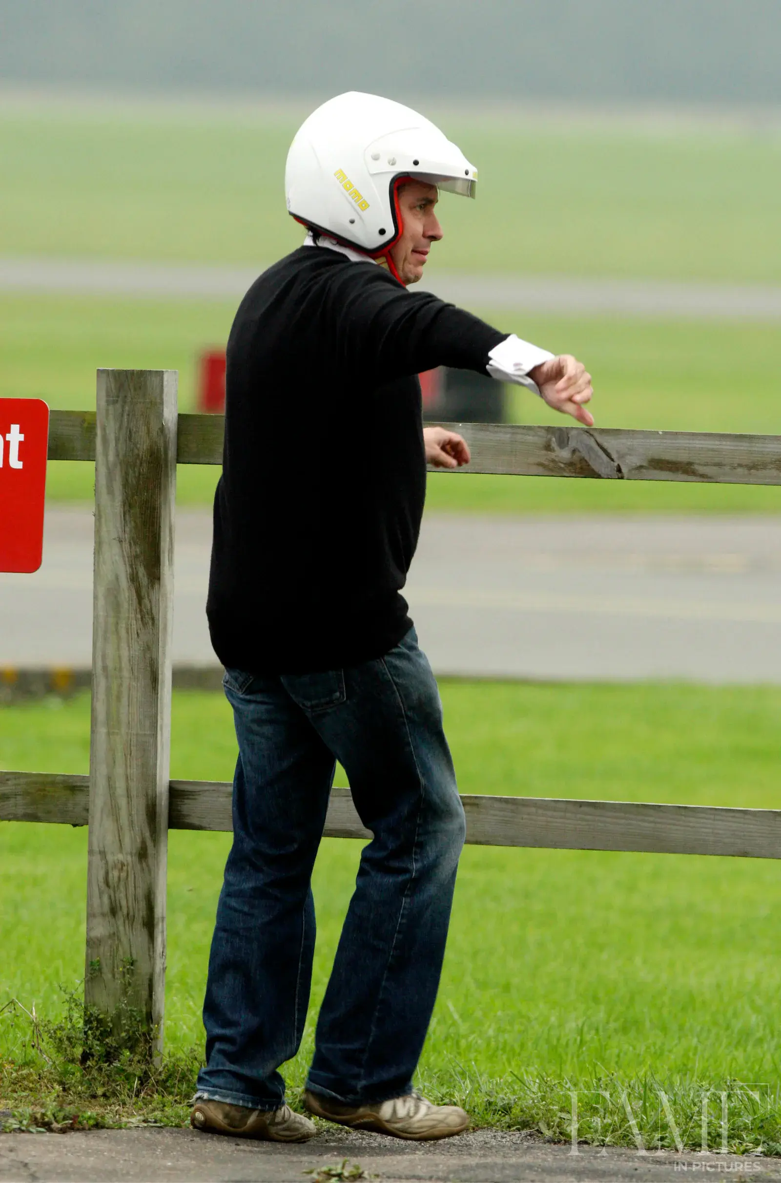 Jools Holland in a white helmet, dark jumper and jeans extending an arm beside a wooden fence