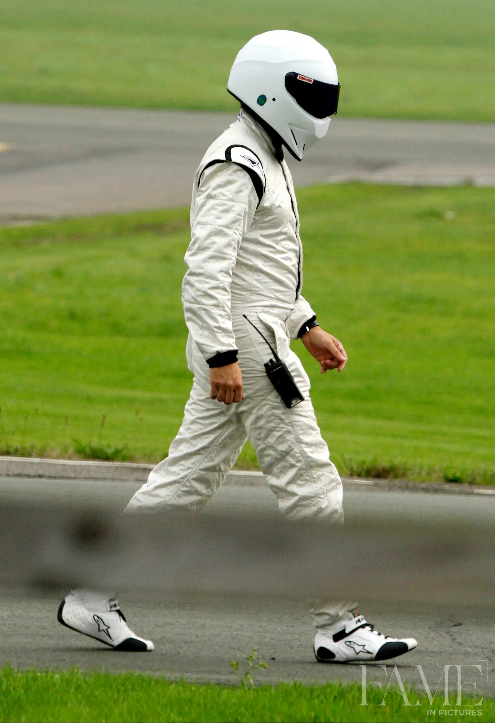 An unidentified driver in white racing gear and a closed-visor helmet walking beside the grass