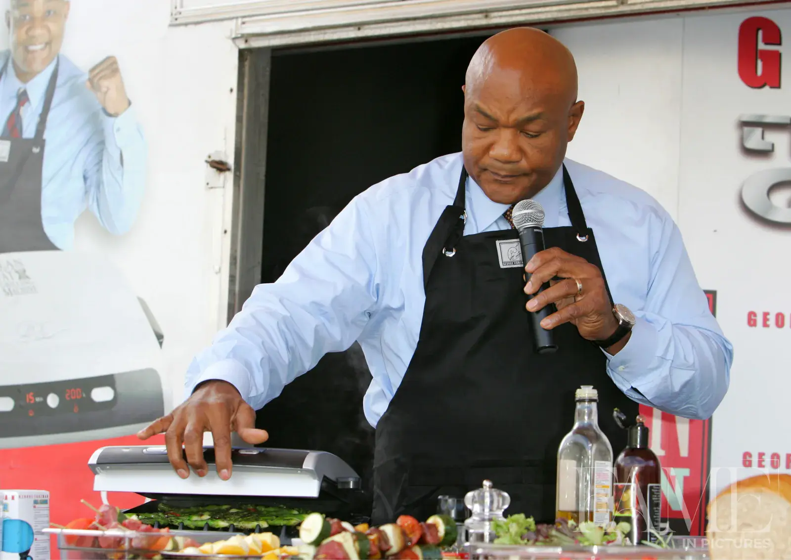 George Foreman demonstrating a grill while holding a microphone, wearing a blue shirt and black apron