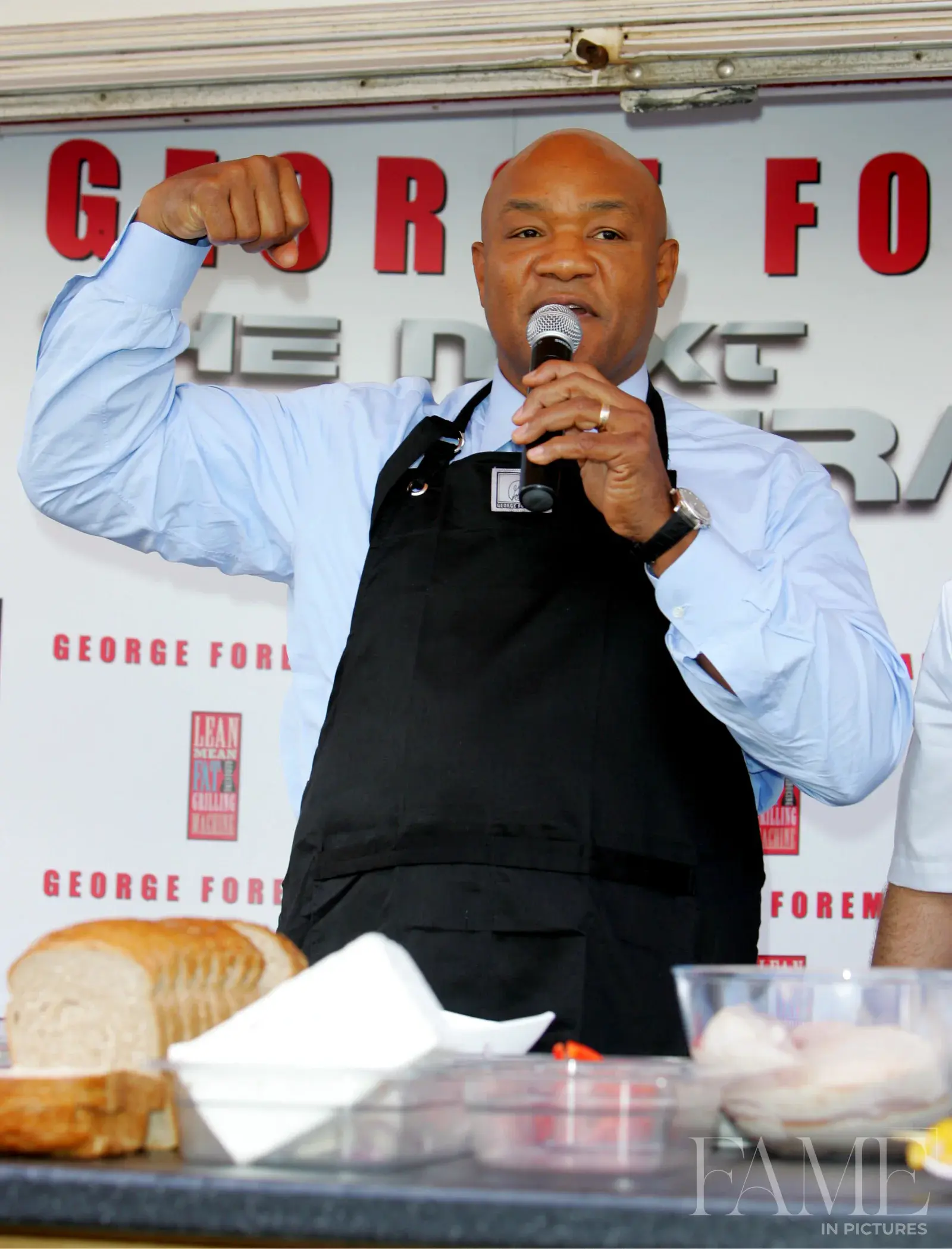 George Foreman raising an arm and holding a microphone behind the demonstration table
