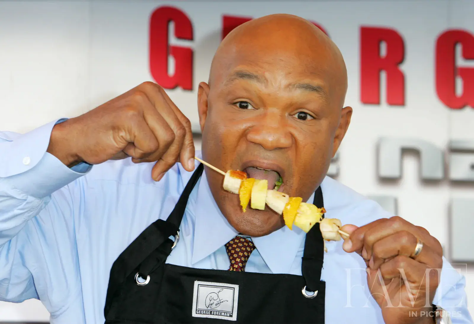 George Foreman tasting food from a skewer while wearing a black apron over a pale blue shirt