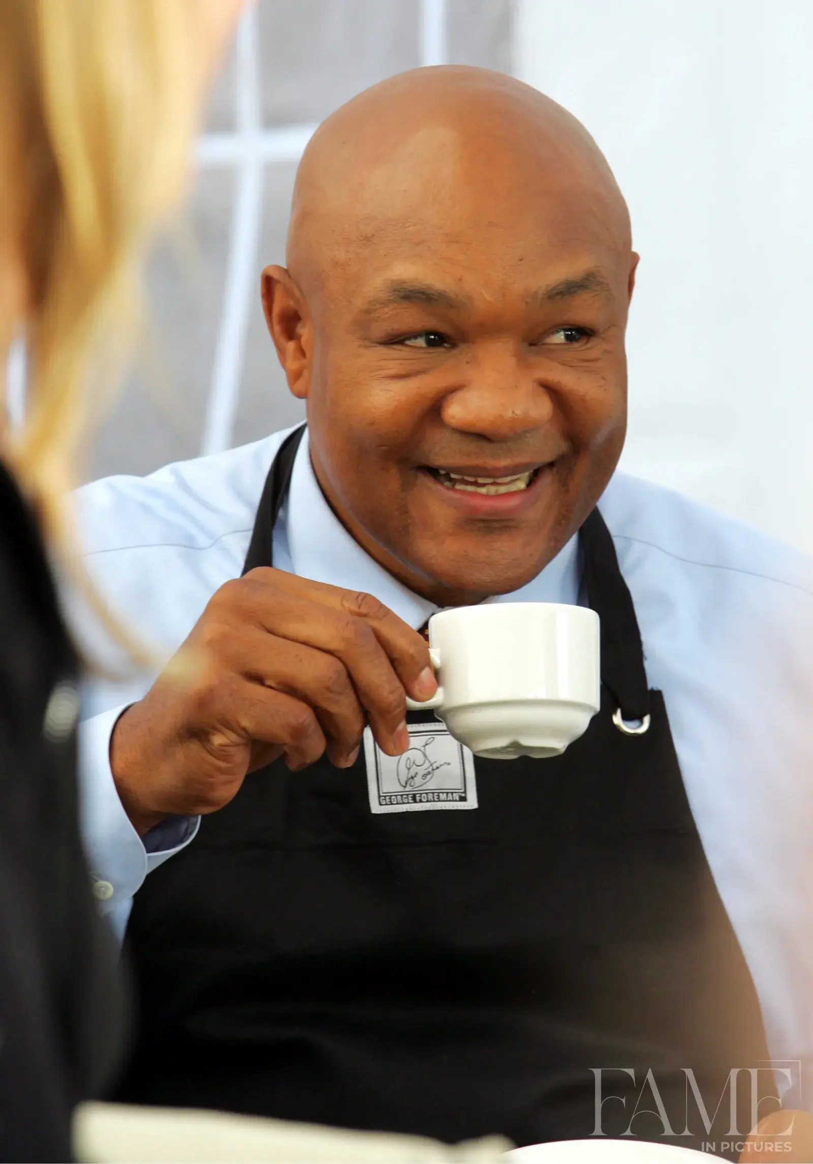 George Foreman holding a small white cup and smiling, wearing a black apron and patterned tie