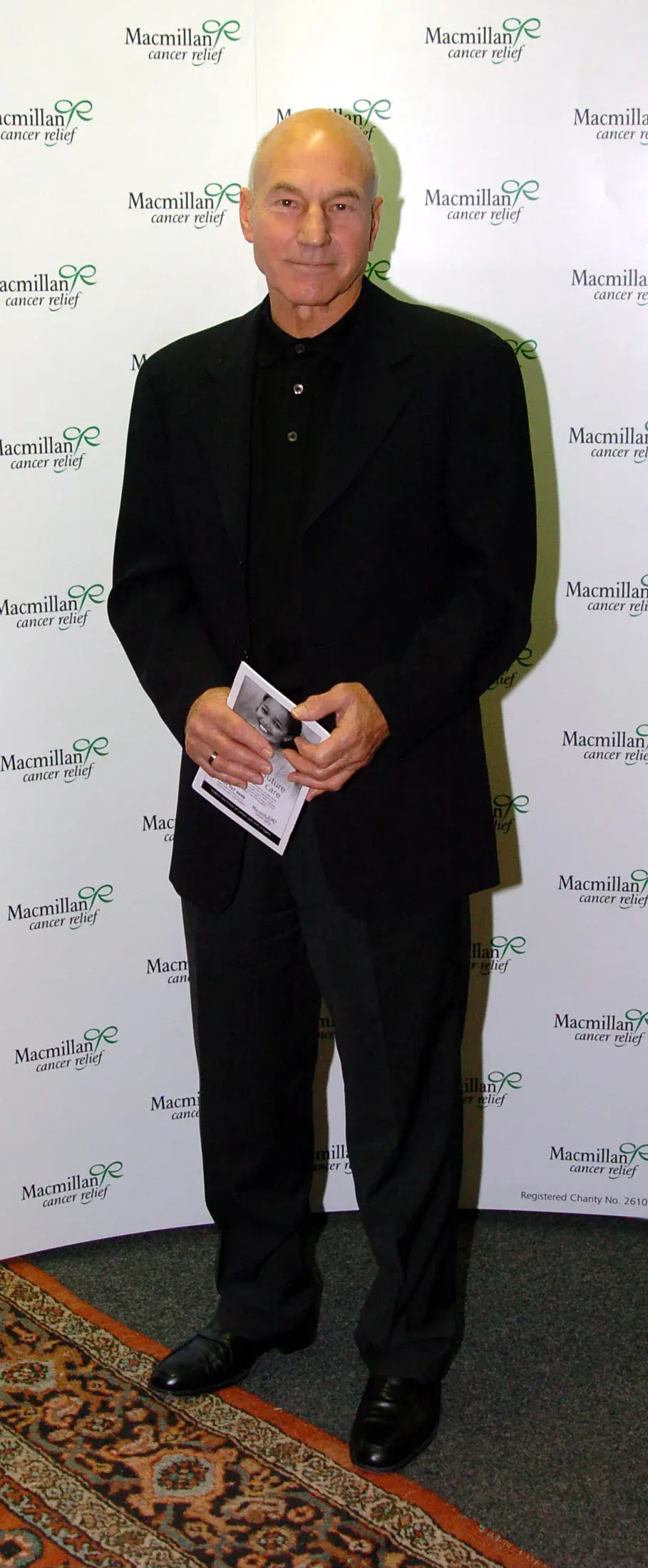 Patrick Stewart in a dark suit holding a programme in front of a Macmillan charity backdrop